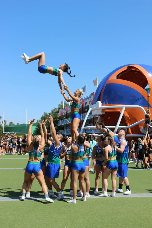 a group of cheerleaders performing a stunt on a field, by John Luke, dreamworld, orange and teal color, clear blue skies, camps in the background