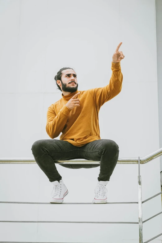 a man sitting on a railing pointing at something, by Ismail Acar, he is wearing a brown sweater, performance, riyahd cassiem, on a gray background