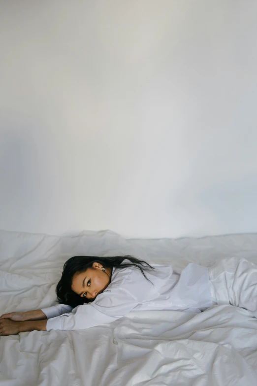 a woman laying on top of a bed covered in white sheets, minimalism, wearing a white sweater, female with long black hair, sad vibe, wearing pajamas