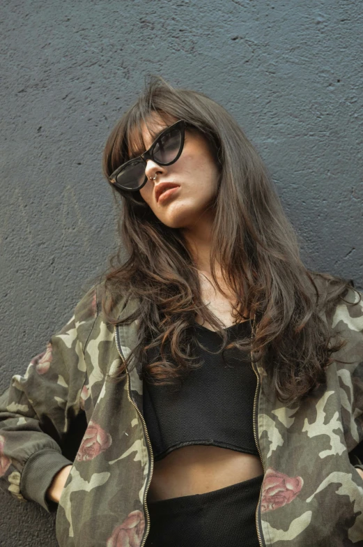 a woman leaning against a wall with her hands on her hips, an album cover, inspired by Elsa Bleda, trending on unsplash, photorealism, fashion model in sunglasses, long dark hair with bangs, wearing camo, headshot profile picture