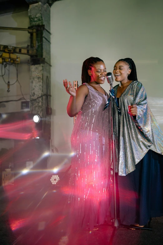 a couple of women standing next to each other, an album cover, by Chinwe Chukwuogo-Roy, pexels, happening, singing at a opera house, industrial party, sparkling light, geometric backdrop; led