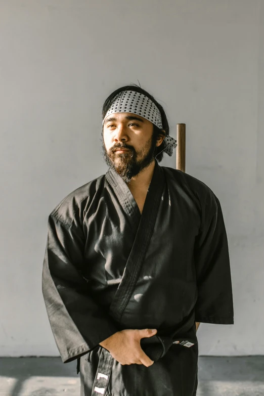 a man in a black kimono standing in front of a white wall, wearing a bandana and chain, lee madgwick & liam wong, on black background, with a beard and a black shirt