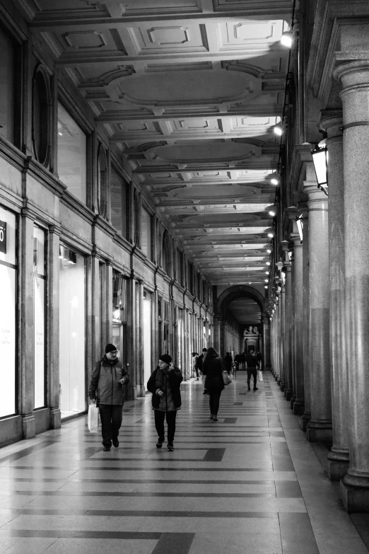 a black and white photo of people walking down a hallway, a black and white photo, flickr, neoclassicism, lots of shops, style of paolo parente, colonnade, late evening