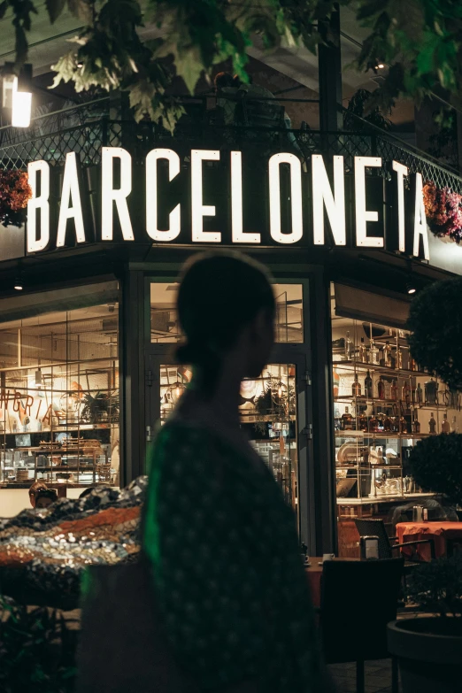 a woman walking in front of a store at night, by Tomàs Barceló, pexels contest winner, baroque, fc barcelona, bright signage, with a city in the background, overview