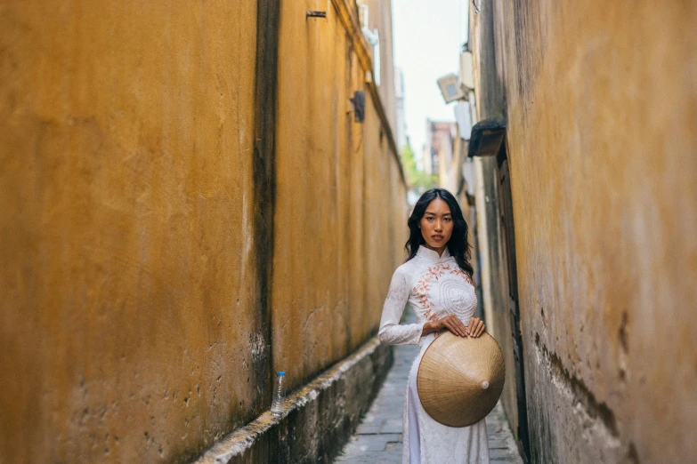 a woman in a white dress is walking down a narrow alleyway, inspired by Ruth Jên, pexels contest winner, ao dai, wearing a round helmet, tan, posing for a picture