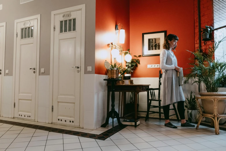 a woman standing next to a table in a room, by Emma Andijewska, pexels contest winner, art nouveau, doctors office, gray and orange colours, people walking around, leaning on door