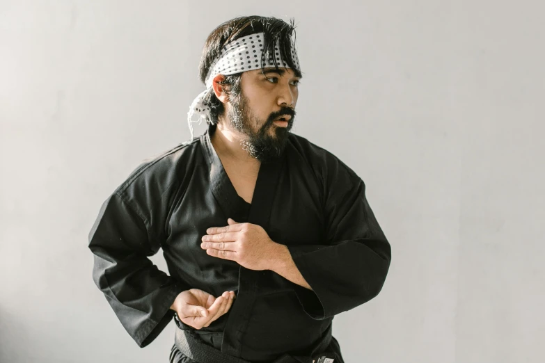 a man in a black kimono standing in front of a white wall, fighting stance, wearing a bandana and chain, matt berry, ash thorp khyzyl saleem