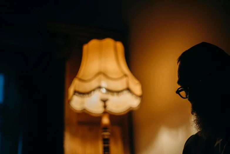 a man standing in front of a lamp in a dark room, inspired by George Jamesone, trending on unsplash, arts and crafts movement, man with glasses, woman silhouette, soft warm light, in a dusty victorian home