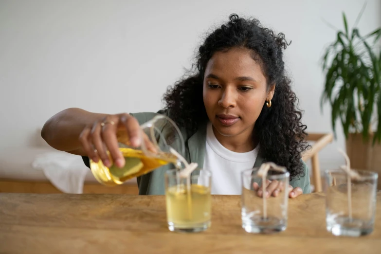 a woman sitting at a table pouring something into a glass, by Jessie Algie, pexels contest winner, manuka, yellow, riyahd cassiem, beakers full of liquid