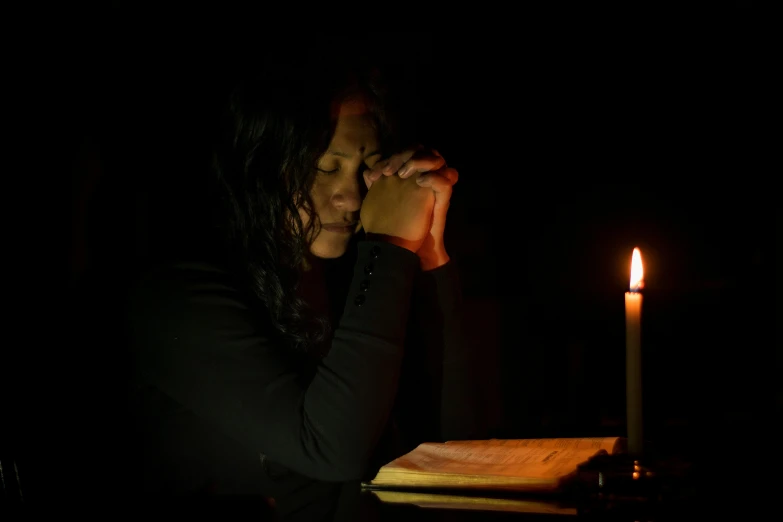 a woman sitting at a table with a candle in front of her, praying, holding electricity, in a black room, staff