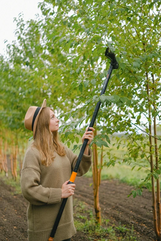 a woman that is standing in the dirt with a pole, with a tall tree, full device, gardening, over-the-shoulder shot