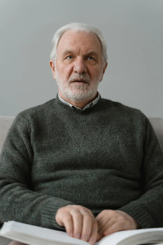 a man sitting on a couch reading a book, inspired by Karl Matzek, some grey hair in beard, looking defiantly at the camera, wearing a sweater, promo image