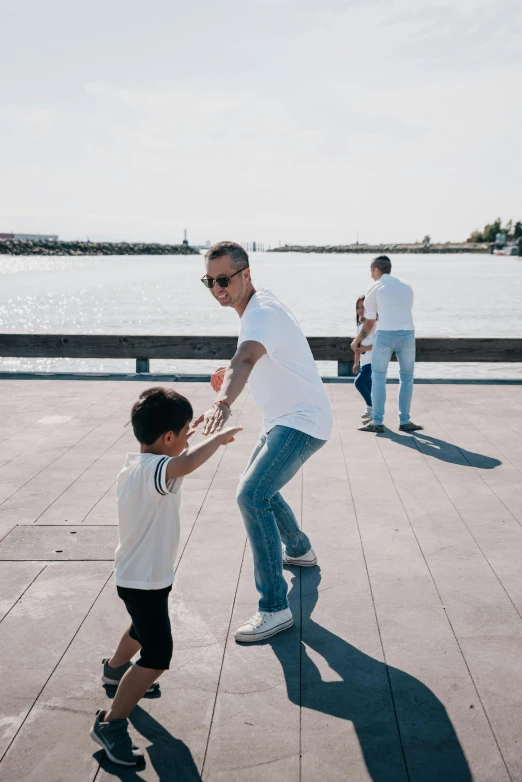 a man riding a skateboard next to a little boy, pexels contest winner, happening, near a jetty, dancing with each other, white shirt and blue jeans, i'm dad