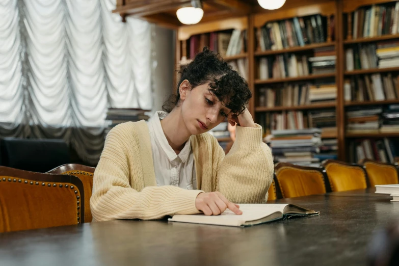 a woman sitting at a table reading a book, pexels, academic art, disappointed, background image, queer woman, dusty library