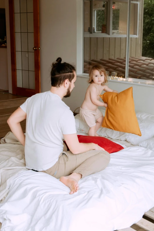 a man sitting on top of a bed next to a baby, pexels contest winner, light beige pillows, fighting, standing, 4yr old