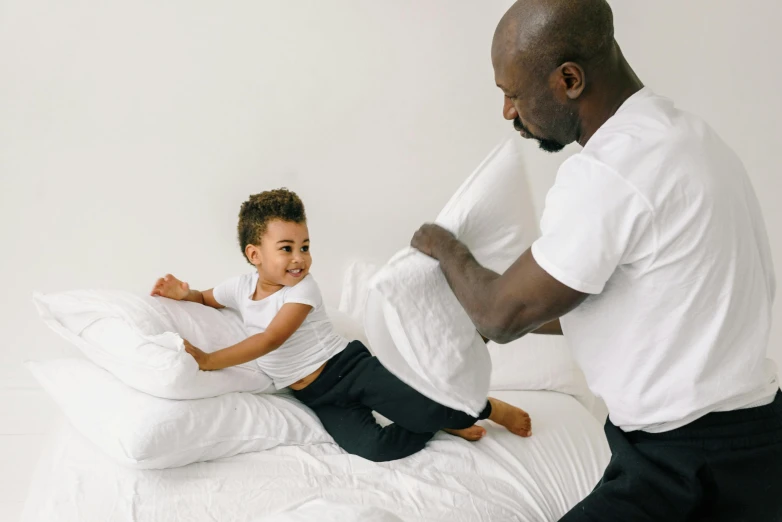 a man and a little girl sitting on a bed, wearing a tee shirt and combats, white pillows, thumbnail, black man