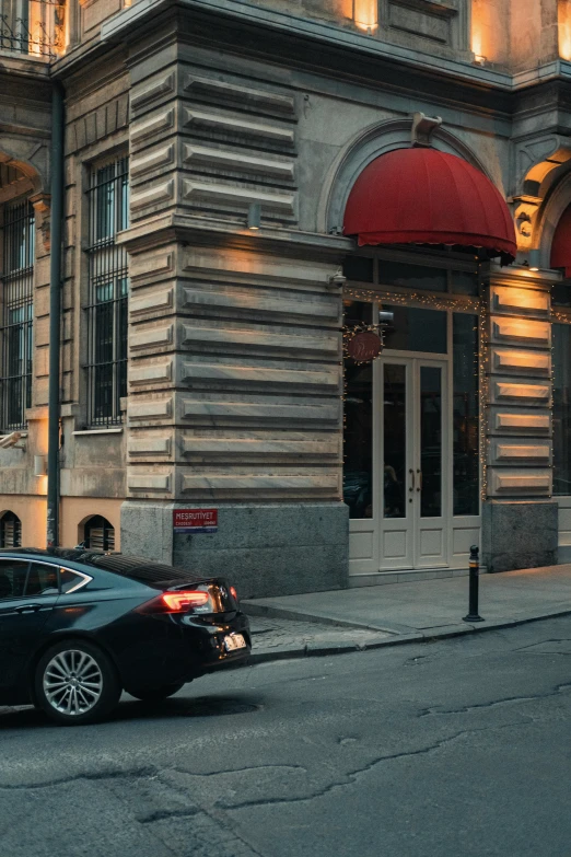 a black car driving down a street next to a tall building, by Géza Mészöly, pexels contest winner, art nouveau, expensive restaurant, tall arched stone doorways, payne's grey and venetian red, summer evening