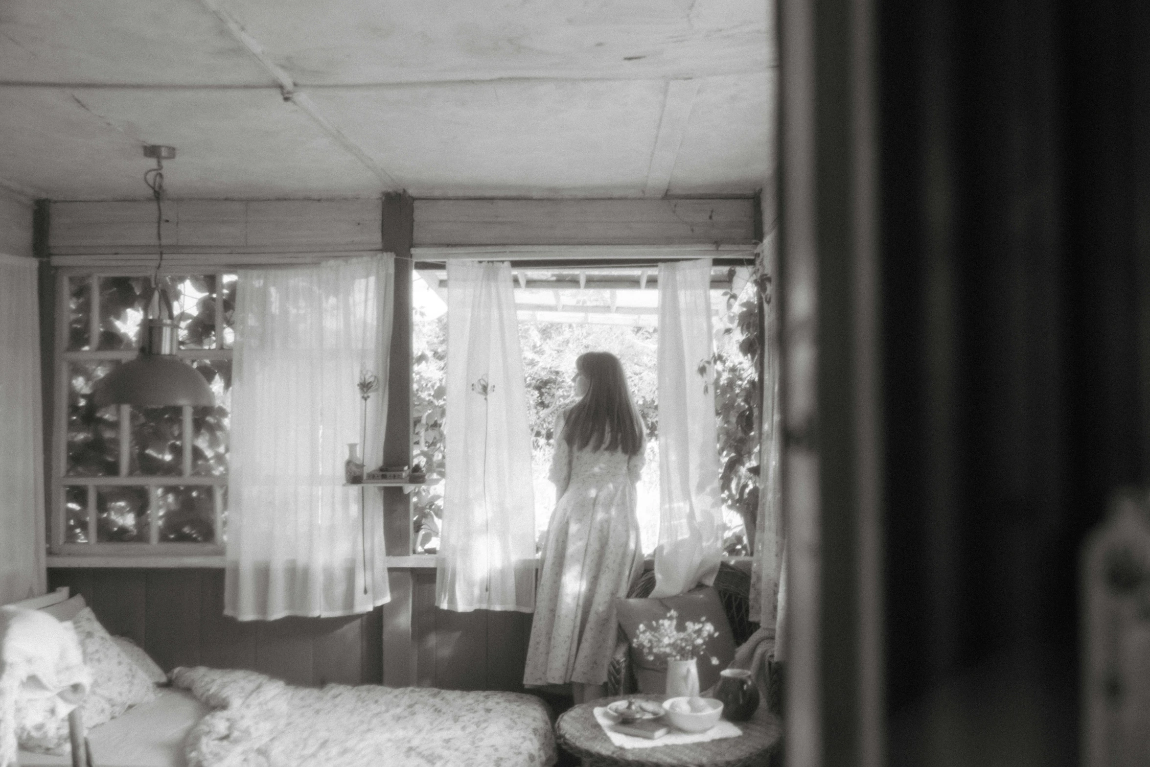 a black and white photo of a woman looking out a window, a black and white photo, inspired by Nell Dorr, romanticism, cottagecore hippie, nico wearing a white dress, bedroom, medium format film photography