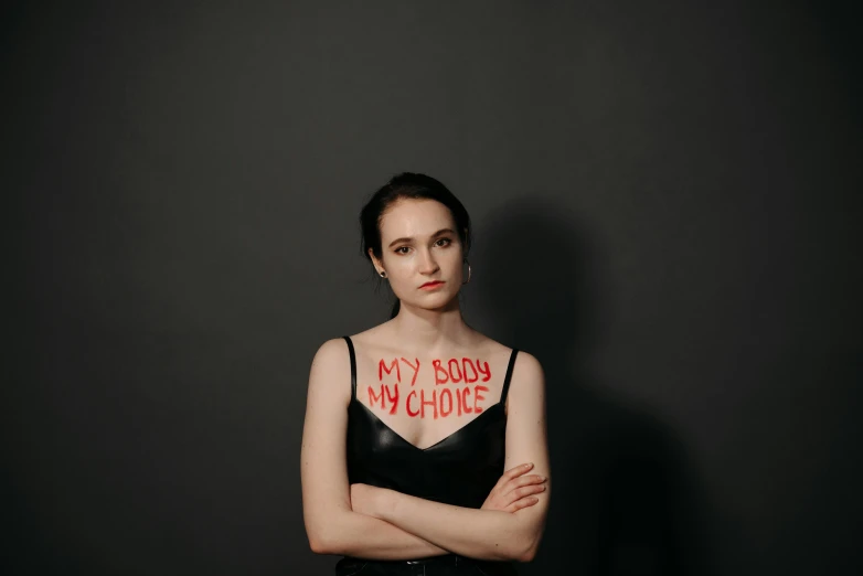 a woman in a black dress with red writing on her chest, by Emma Andijewska, pexels, sots art, winona ryder, woman holding sign, 19-year-old girl, semi-transparent