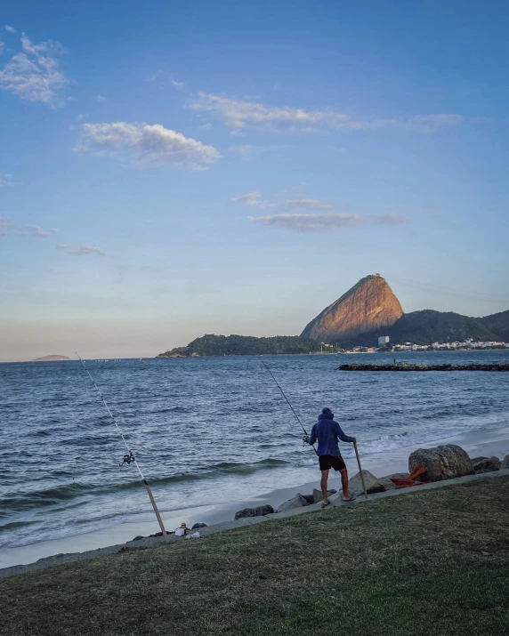 a man standing on top of a beach next to a body of water, by Elsa Bleda, pexels contest winner, plein air, cristo redentor, fishing, lgbtq, standing in front of a mountain