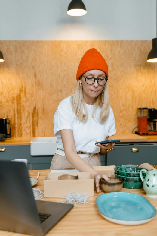 a woman sitting at a kitchen table in front of a laptop, a portrait, trending on pexels, arbeitsrat für kunst, beanie, sitting on a store shelf, low quality photo, square