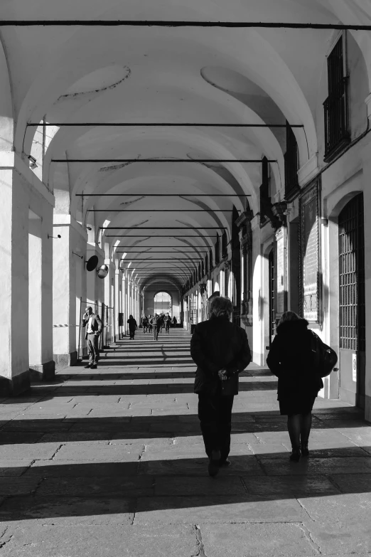 a couple of people walking down a hallway, inspired by Henri Cartier-Bresson, minimalism, lots of roman arches, in empty!!!! legnica, some stalls, naples