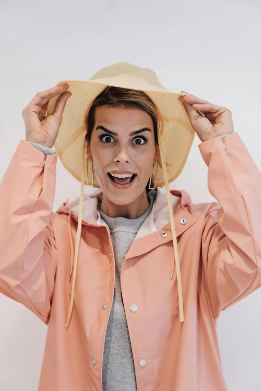 a woman holding a hat on top of her head, pexels contest winner, girl in raincoat, surprised frown, in shades of peach, winking at the camera