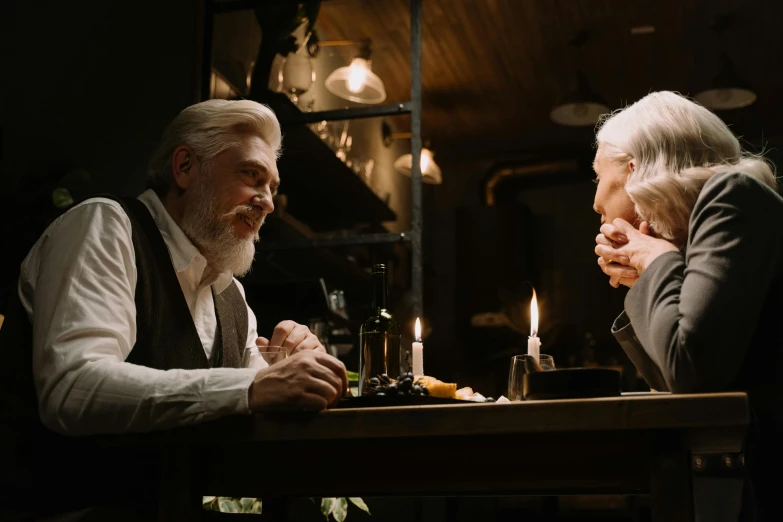 a man and a woman sitting at a table, by Emma Andijewska, pexels contest winner, magical realism, white hair and beard, old movie scene, candlelit, ( ( theatrical ) )