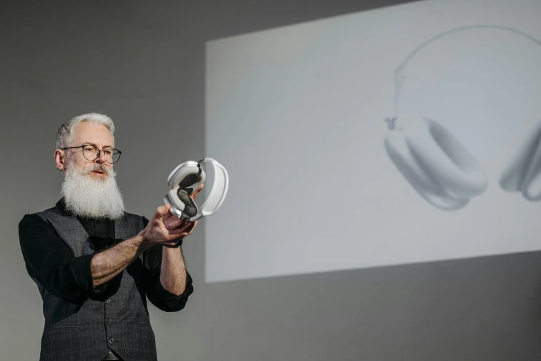 a man standing in front of a projector screen holding a pair of headphones, a hologram, by Haukur Halldórsson, long white hair and white beard, holding helmet, 3 d print, vessels