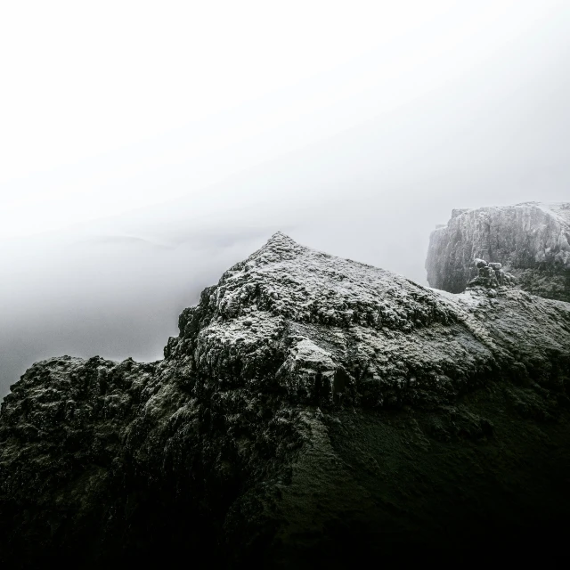 a person standing on top of a snow covered mountain, a black and white photo, pexels contest winner, romanticism, uneven dense fog, faroe island, dark rocks, shot from a drone