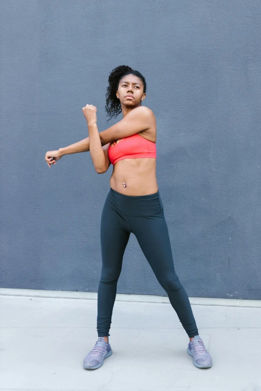 a woman standing in front of a gray wall holding a tennis racquet, happening, boxing stance, wearing a crop top, arms stretched out, alexis franklin