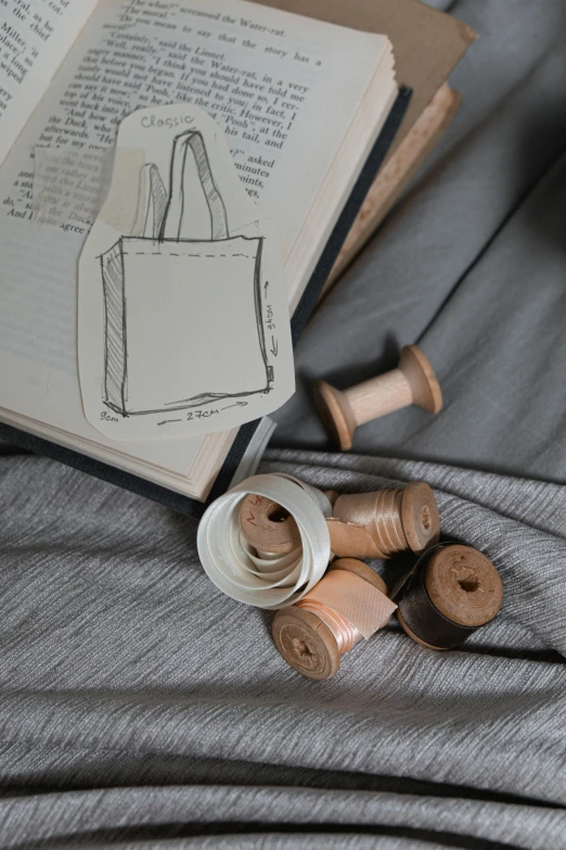 an open book sitting on top of a bed next to spools of thread, a sketch, featured on pinterest, bags, sustainable materials, detail shot, grey robes