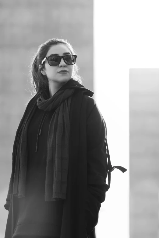 a woman standing in front of a tall building, a black and white photo, sunglasses and a scarf, wearing a black hoodie, mina petrovic, pondering