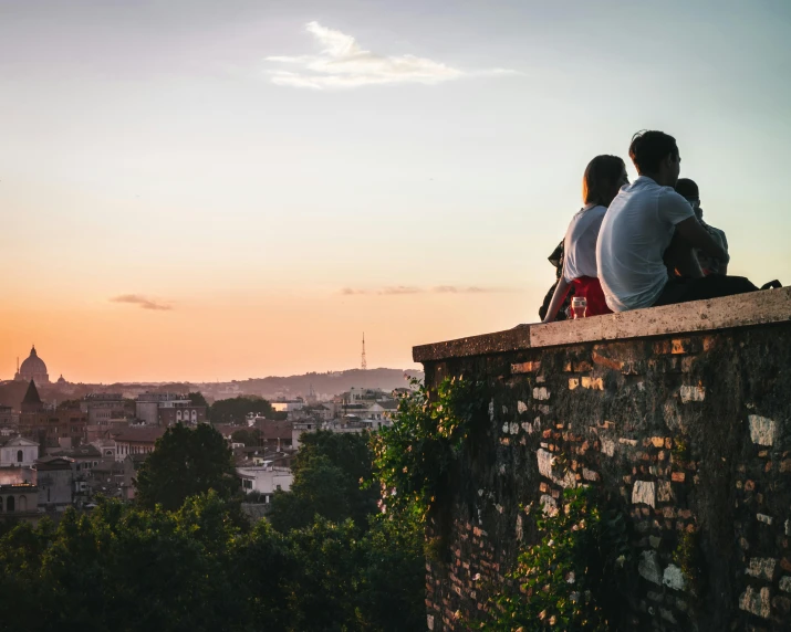 two people sitting on a wall overlooking a city, by Lucia Peka, pexels contest winner, summer evening, roma, family friendly, city wall