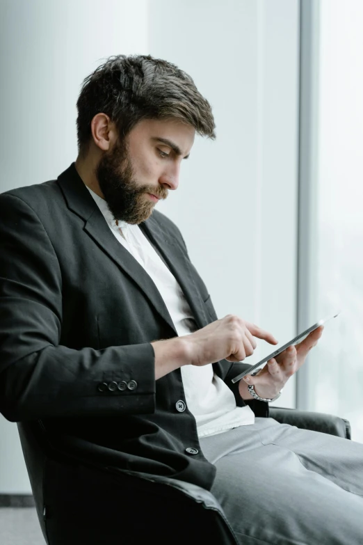 a man sitting in a chair looking at a cell phone, with a beard and a black jacket, it specialist, wearing a white button up shirt, using a magical tablet