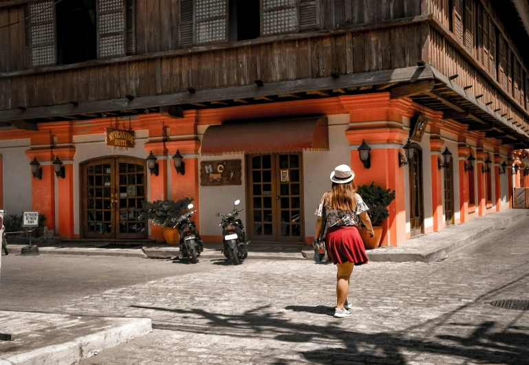 a woman is walking down a cobblestone street, pexels contest winner, philippines, red building, dressed in a top and shorts, caracter with brown hat