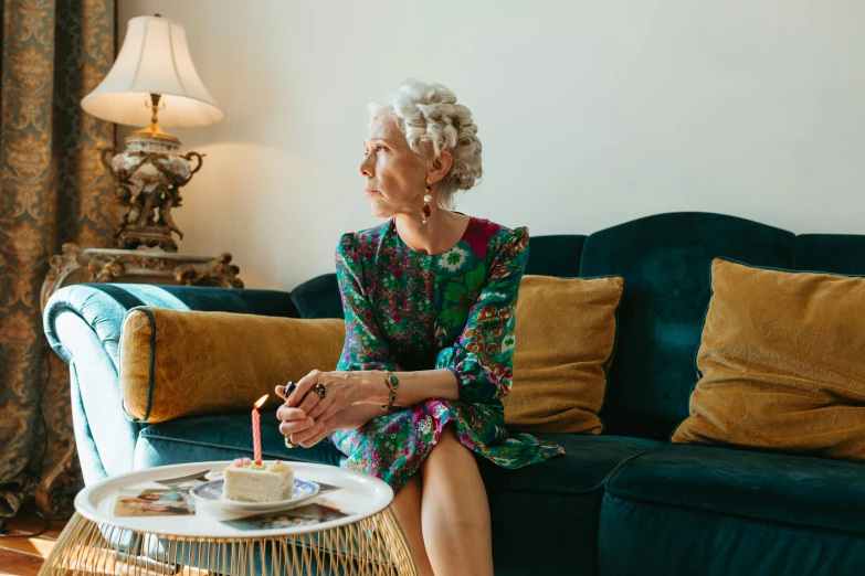 a woman sitting on a couch in a living room, by Lucette Barker, pexels, maximalism, holding a birthday cake, gray haired, she wears a dark green dress, pondering