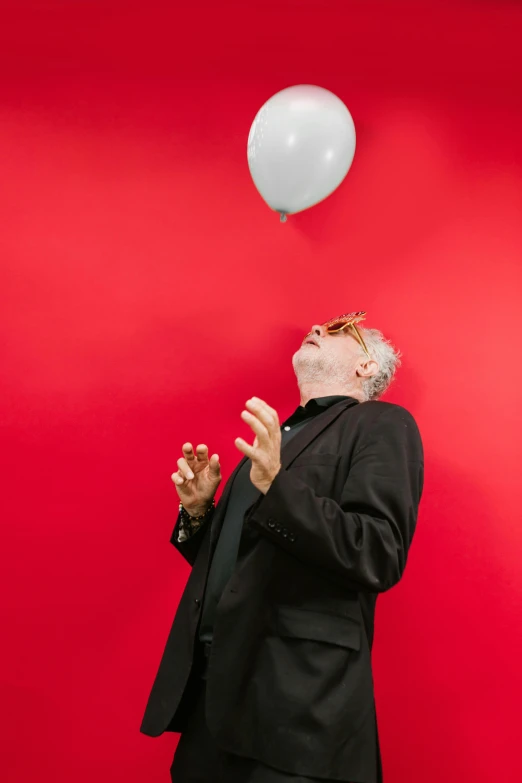 a man standing in front of a red wall with a white balloon, inspired by Shōzō Shimamoto, a silver haired mad, screaming into air, silver small small small glasses, ignant