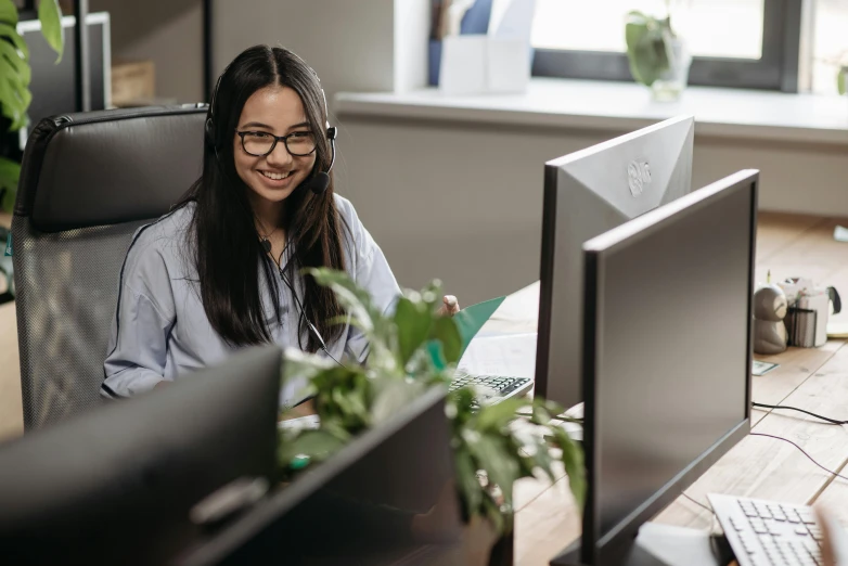 a woman sitting at a desk in front of two computer monitors, trending on pexels, hurufiyya, smiling down from above, in an call centre office, nvidia promotional image, darren quach