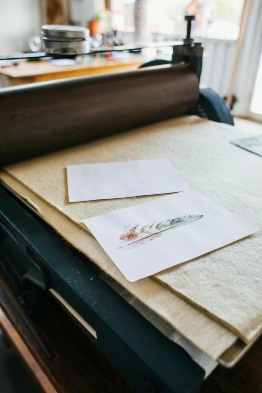 a sheet of paper sitting on top of a machine, a silk screen, by Jessie Algie, unsplash, private press, real feather, ilustration, restoration, on a white table