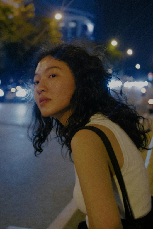 a woman sitting on a bench in a city at night, inspired by Feng Zhu, trending on unsplash, photorealism, her face framed with curls, film still from the movie, jaeyeon nam, pictured from the shoulders up