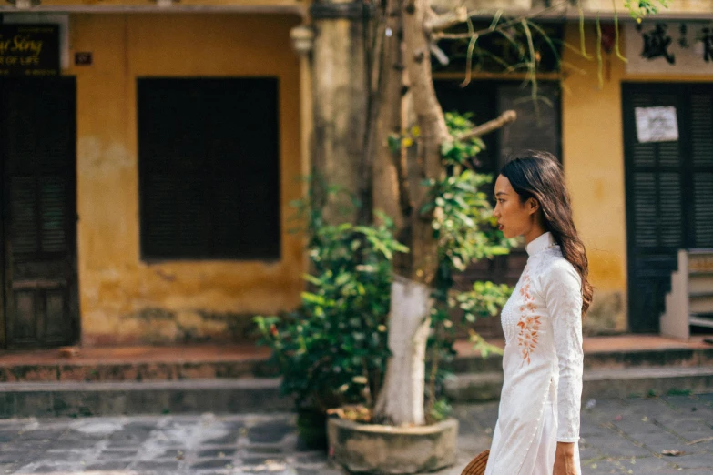 a woman in a white dress walking down a street, a photo, inspired by Ruth Jên, pexels contest winner, happening, wearing a silk kurta, vietnamese woman, ancient garden behind her, tan