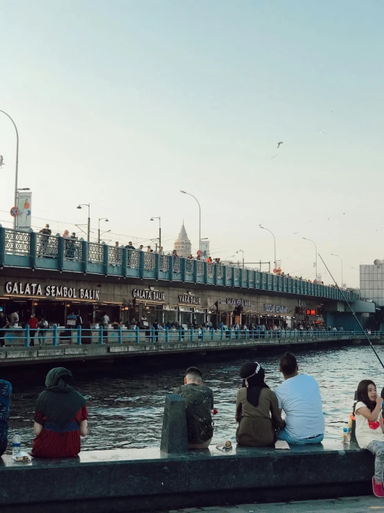 a group of people sitting next to a body of water, graffiti, on a bridge, turkish and russian, fish seafood markets, at golden hour