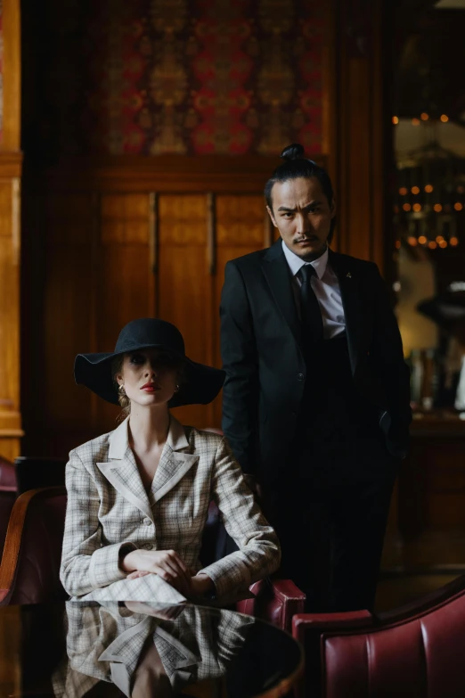 a woman sitting in a chair next to a man in a suit, unsplash, art nouveau, joe taslim, a suited man in a hat, dramatic film still, [ theatrical ]