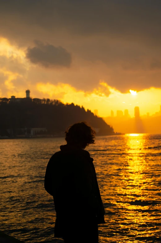 a person standing next to a body of water at sunset, by Nabil Kanso, istanbul, skyline showing, head turned, yellow