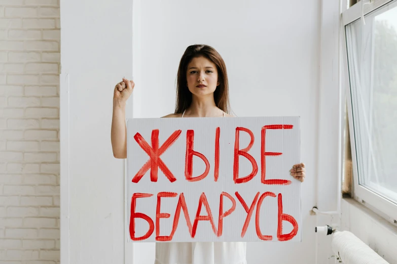 a woman holding a sign in front of a window, a poster, pixabay, feminist art, president of belorussia, on a white table, background image, teenage no