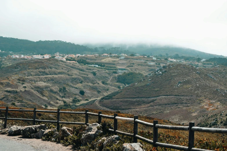 a bench sitting on the side of a road next to a fence, pexels contest winner, les nabis, view of villages, alvaro siza, hollister ranch, in the bottom there a lot of fog