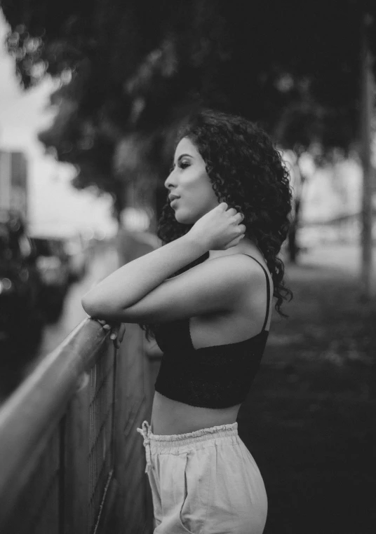 a black and white photo of a woman leaning against a fence, by Alexis Grimou, wearing a black cropped tank top, mixed-race woman, trending photo, thoughtful )