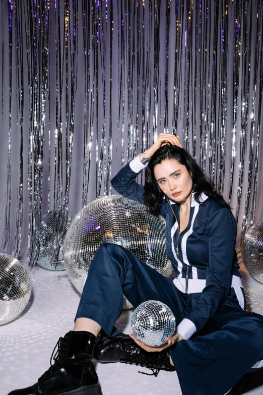 a woman sitting on a bed holding a disco ball, by Julia Pishtar, wearing prison jumpsuit, lilly collins, official store photo, complex background