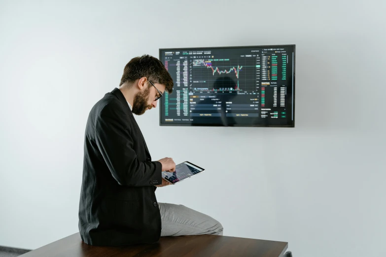 a man sitting on a desk in front of a monitor, a picture, trending on pexels, analytical art, giant crypto vault, a man wearing a black jacket, blackboard in background, large screen